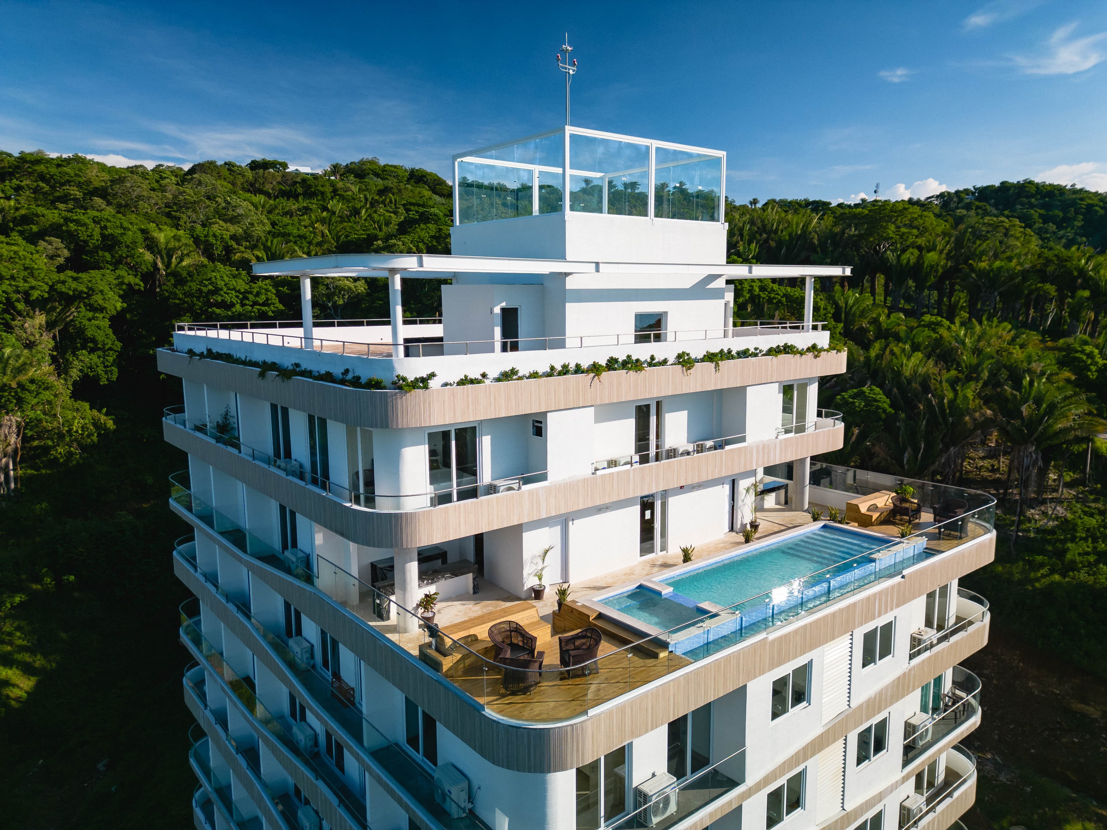 A modern multi-story luxury apartment or condominium building with white exterior walls and large balconies. The top floor features a glass-enclosed area. A long infinity pool is visible on one of the lower balconies, surrounded by wooden decking and outdoor furniture. The building is set against a backdrop of lush green tropical forest.