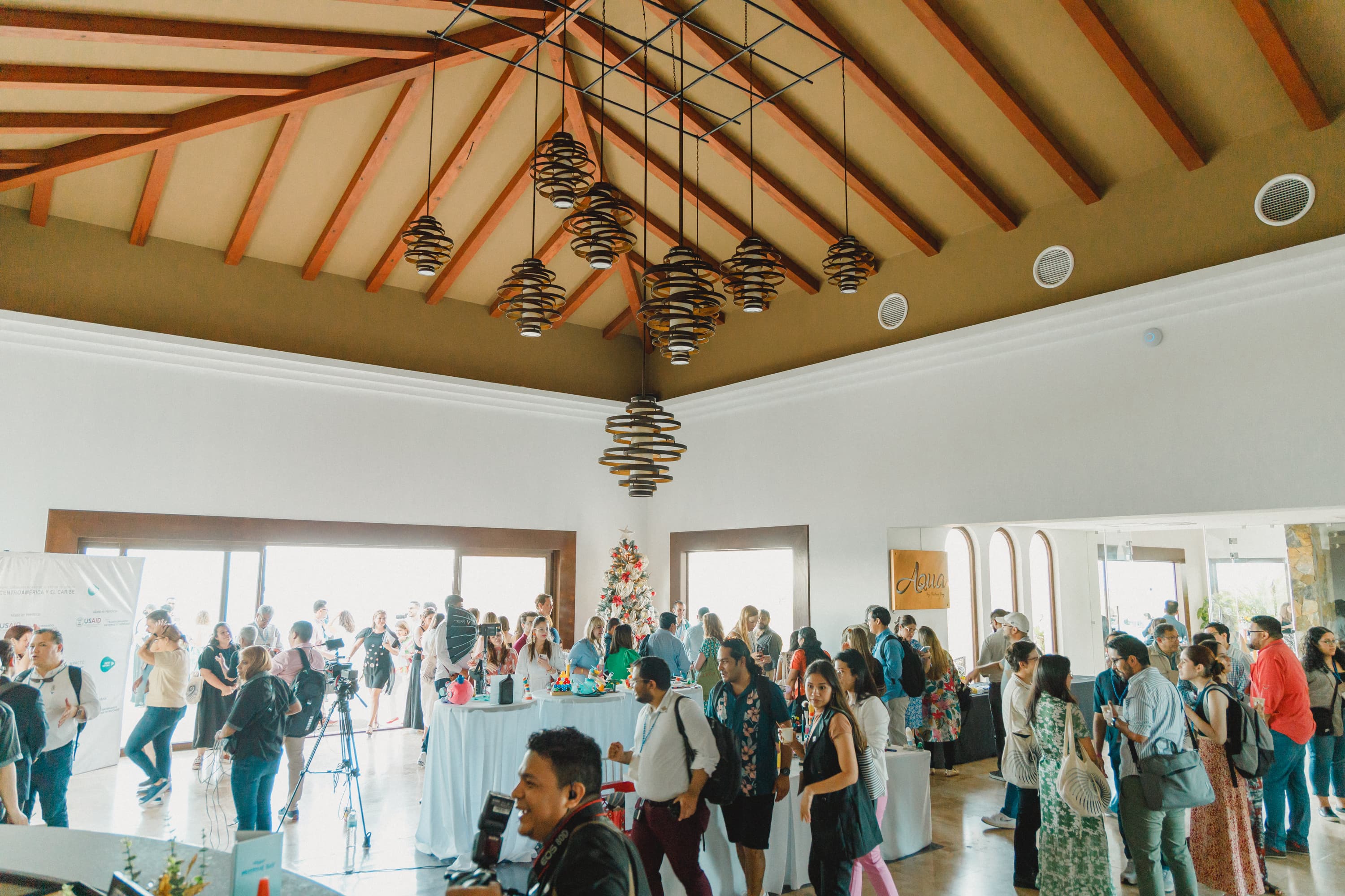 A large group of people gathered in a spacious, well-lit room with high, wooden-beamed ceilings, featuring modern pendant lights. The crowd appears to be mingling around tables with drinks and food, with some individuals using cameras and other recording equipment.